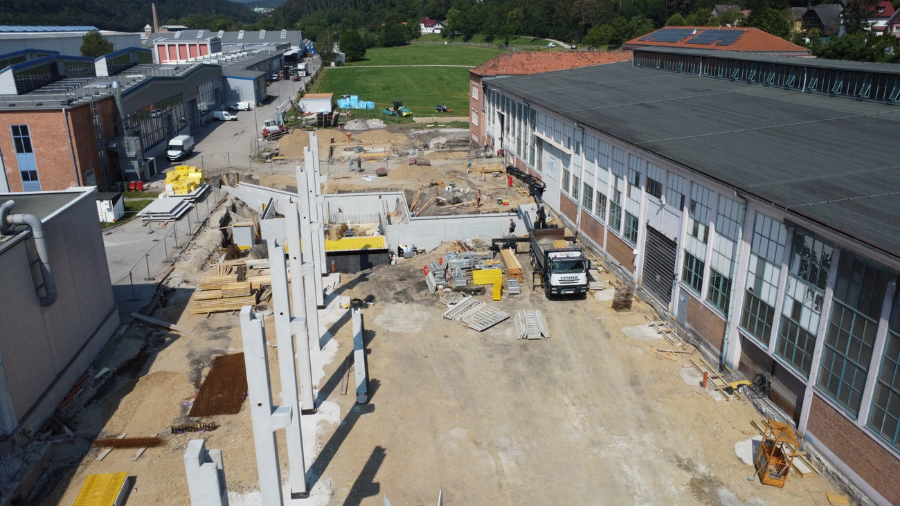 Luftaufnahme einer Baustelle mit Betonpfeilern und Bodenarbeiten neben bestehenden Industriegebäuden.