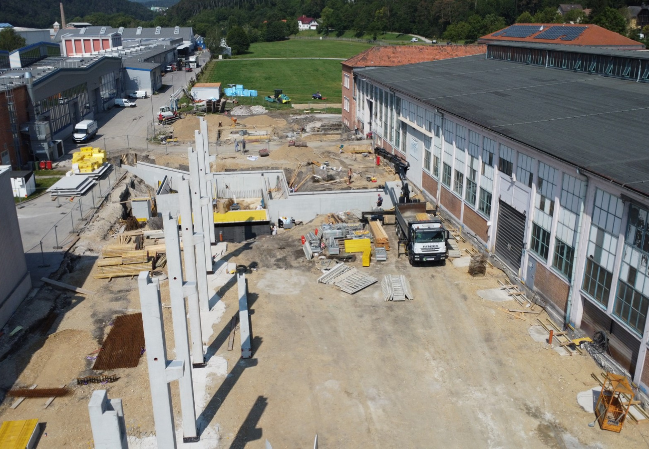 Luftaufnahme einer Baustelle mit Betonpfeilern und Bodenarbeiten neben bestehenden Industriegebäuden.