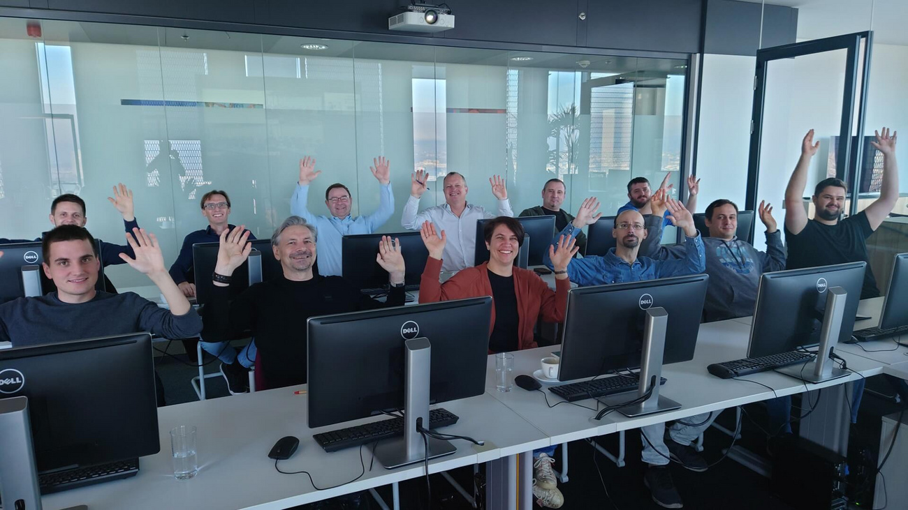 Das Team von Pfnier & Co GmbH während der Schulung zur Nevaris Bausoftware. Eine Gruppe von Mitarbeitern sitzt motiviert an Computerarbeitsplätzen und hebt lächelnd die Hände. Die moderne Schulungsumgebung mit großen Fenstern und Tageslicht ist zu sehen.