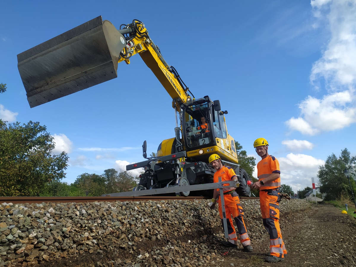 Ein Liebherr Schienenbagger hebt eine Betonplatte, während zwei Arbeiter in Schutzkleidung Vorbereitungen entlang der Schiene treffen.