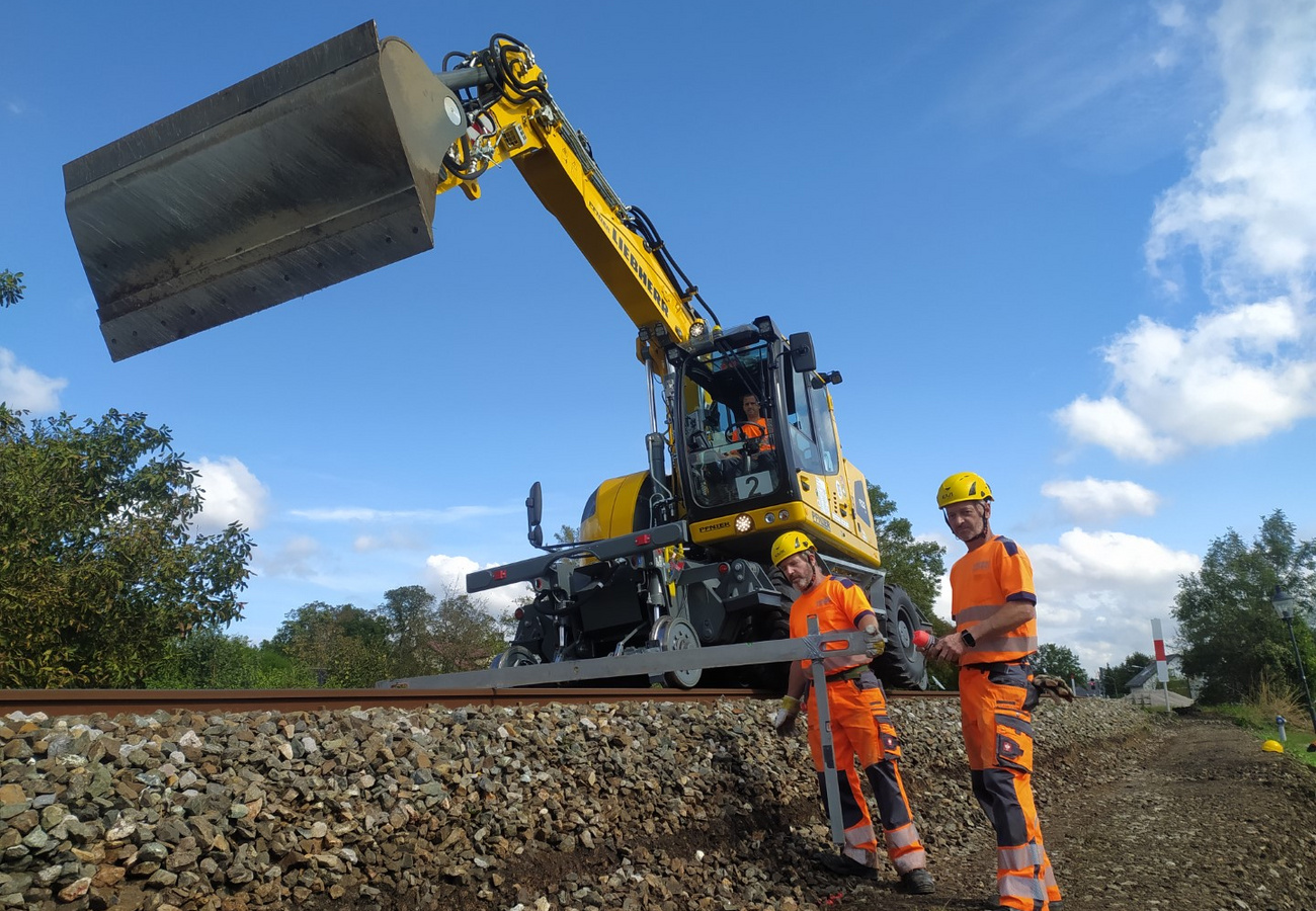 Ein Liebherr Schienenbagger hebt eine Betonplatte, während zwei Arbeiter in Schutzkleidung Vorbereitungen entlang der Schiene treffen.